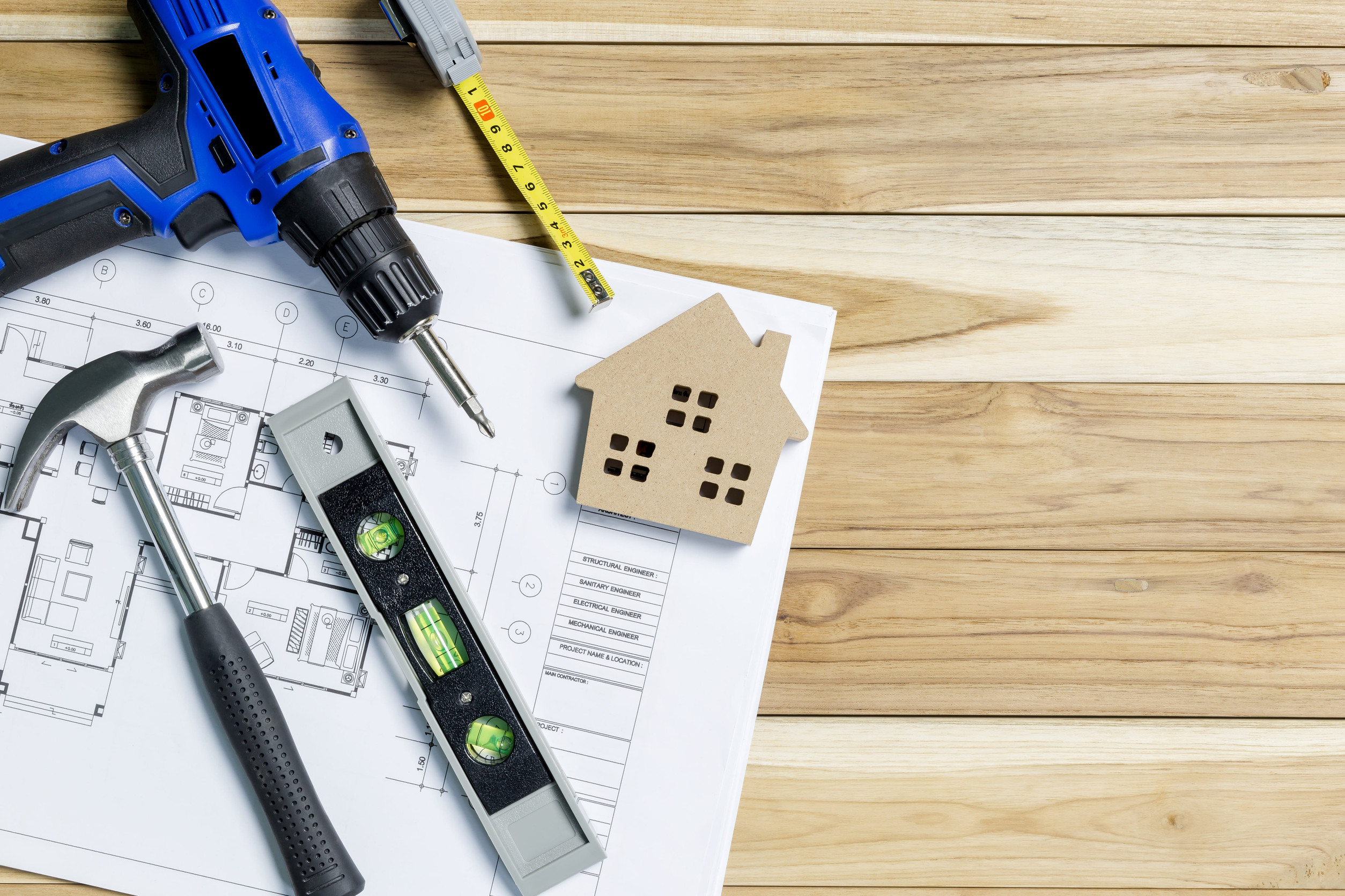 Home renovation tools including a drill, hammer, spirit level, tape measure, and house model placed on architectural floor plans over a wooden surface, representing home improvement, remodeling, or construction planning.